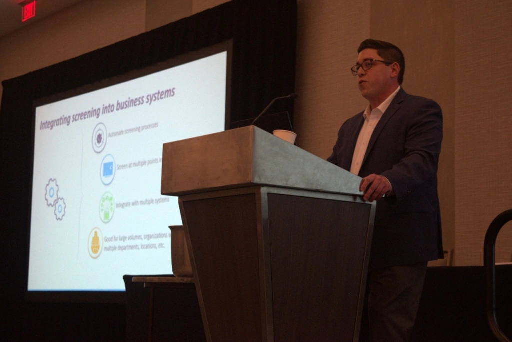 Descartes sales executive Max Elias delivering a presentation at a podium, with a projected slide illustrating how Descartes' integrated denied party screening solution empowers universities to proactively manage export compliance risks.